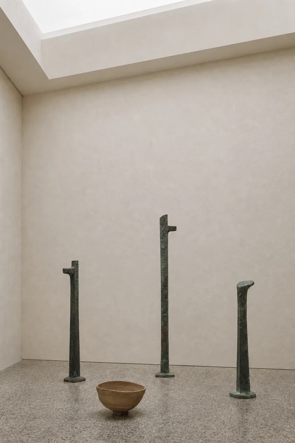 Three slender bronze forms and a clay vessel in a pale gallery
